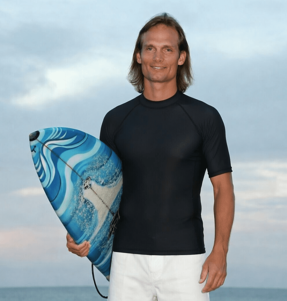 Man standing on a beach holding a blue surfboard with a wave design, wearing a black rash guard and white shorts, with the ocean and sky in the background.