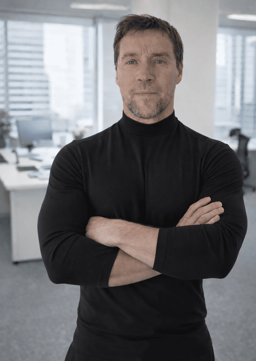 A man in a black turtleneck stands with arms crossed in a modern office with desks, computers, and large windows in the background.